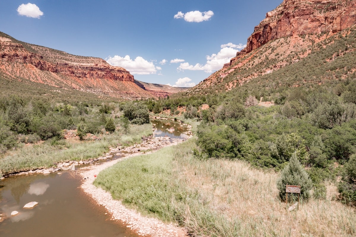 The Dolores River narrows to a trickle with rock banks, flanked by vegetation and red cliffs.